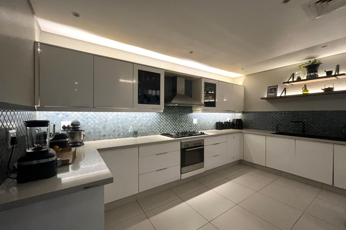 Modern kitchen featuring high-gloss cabinetry, stone countertops, integrated gas hob, stainless steel oven, extractor fan, and decorative tiled backsplash with ambient under-cabinet lighting.