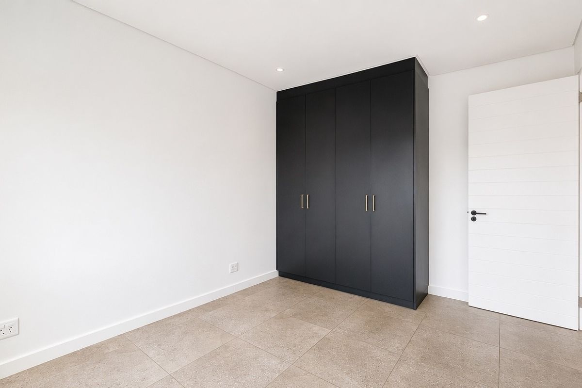 A bright, modern bedroom featuring neutral tiled flooring, crisp white walls, and sleek dark built-in cupboards.