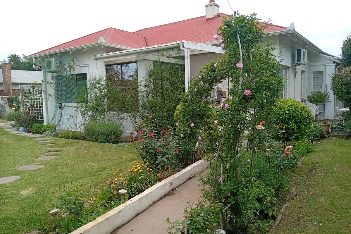 A view of the rear exterior of the house featuring a red metal roof, a covered patio area, and a well-established garden with a concrete pathway.