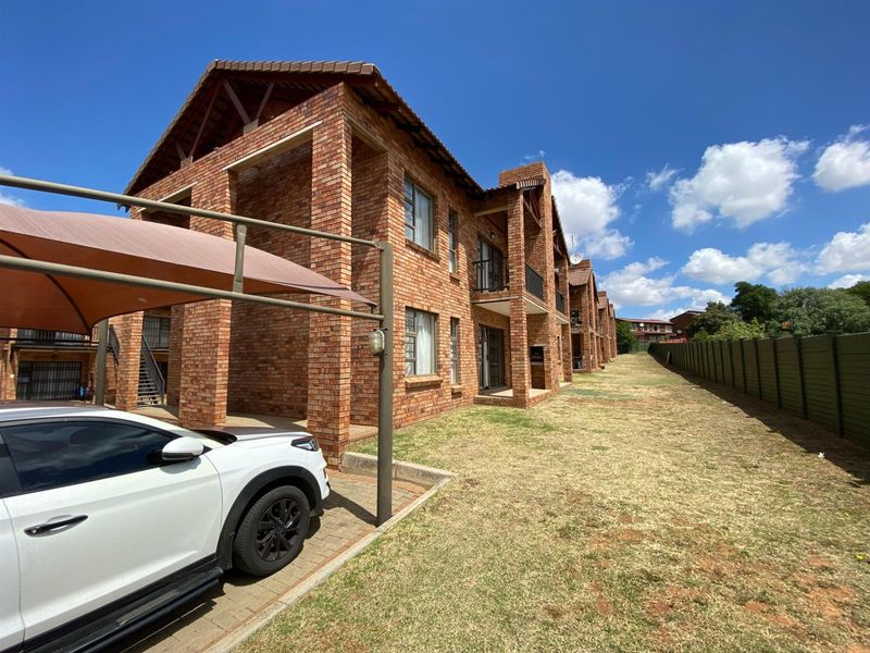 Exterior view of a multi-story face brick apartment complex featuring covered patios, tiled roofing, and a paved parking area with carport structures.
