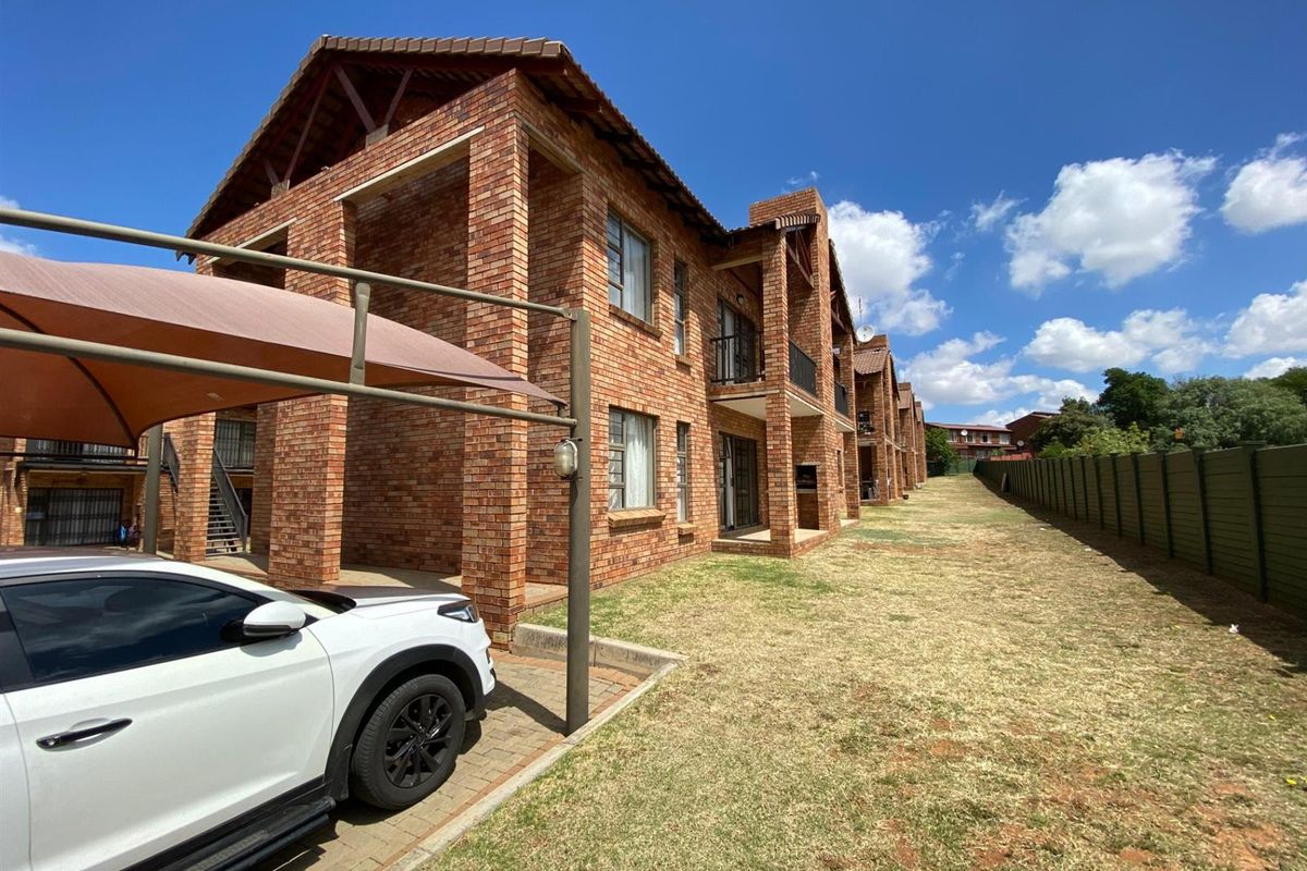 Exterior view of a multi-story face brick apartment complex featuring covered patios, tiled roofing, and a paved parking area with carport structures.