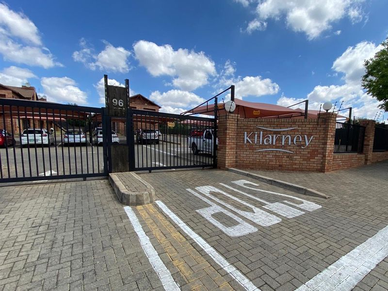 The front entrance of Kilarney complex featuring a secure metal gate, brick boundary wall, and paved driveway with covered parking bays.