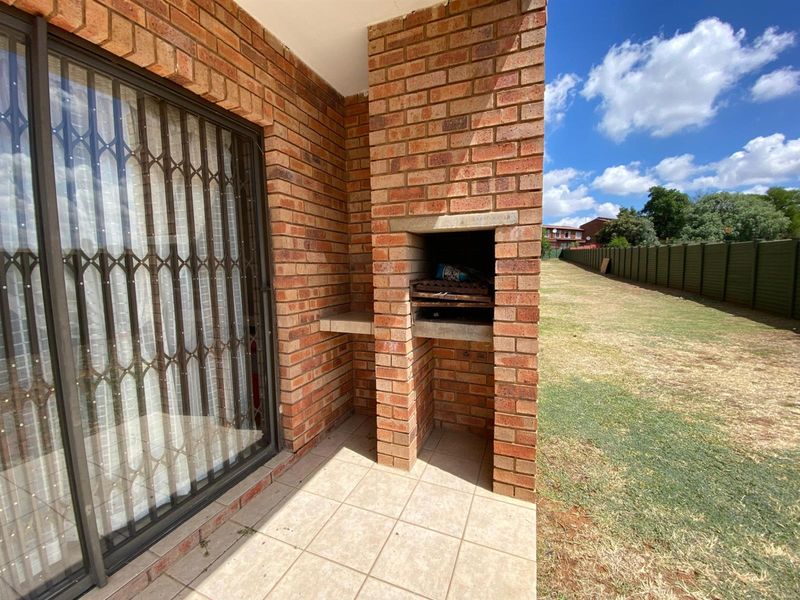 Covered patio area featuring a built-in brick braai, tiled flooring, and a sliding glass door with security gate, overlooking the rear garden.