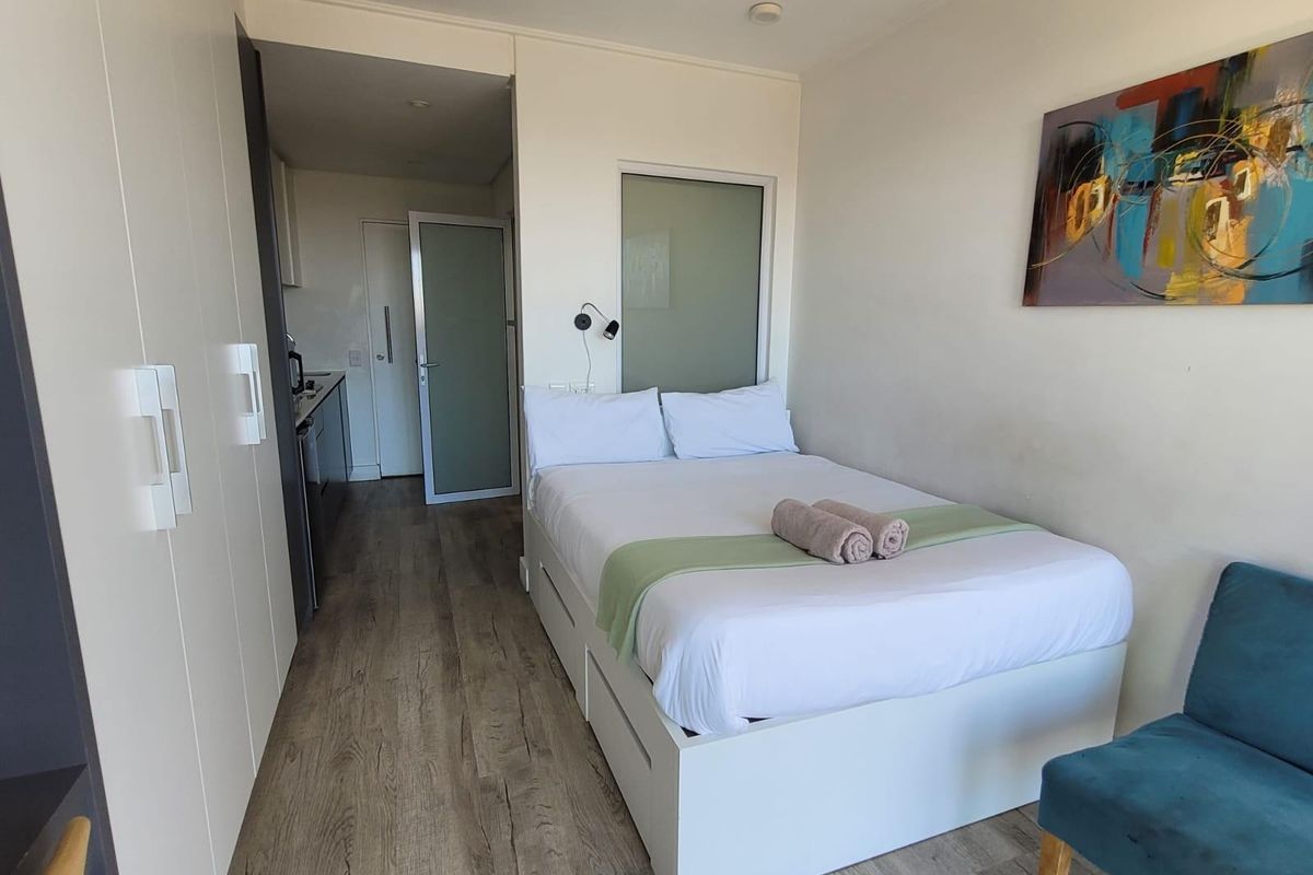 A modern studio bedroom featuring laminate flooring, built-in storage cupboards, and a frosted glass partition, with an adjacent kitchenette area.