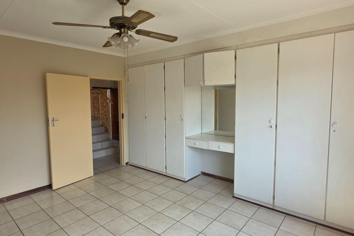 Spacious bedroom featuring built-in cupboards with an integrated vanity, tiled flooring, and a ceiling fan, with an adjacent bathroom and hallway access.