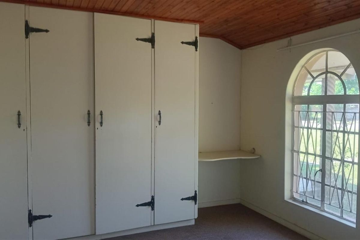 A bedroom featuring built-in storage cupboards, a small integrated desk shelf, and an arched window with security bars and wood-paneled ceiling.