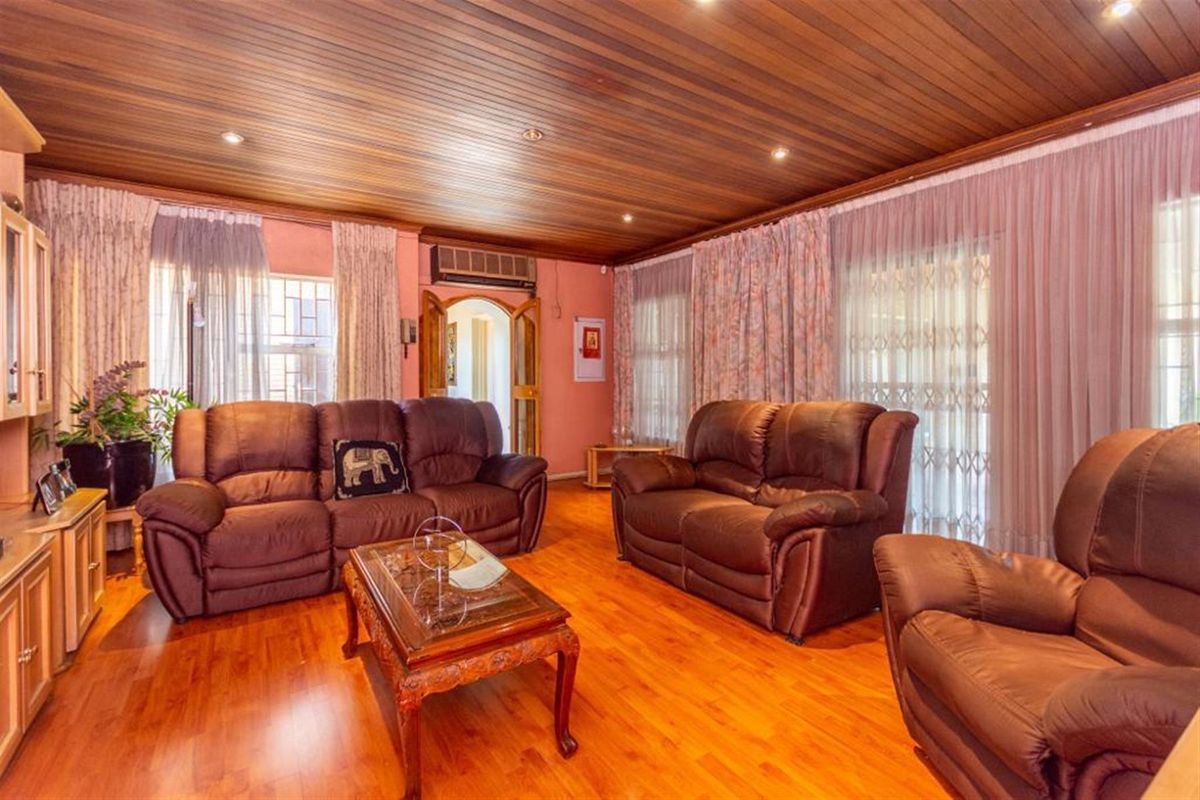 A spacious living room featuring warm timber-paneled ceilings, laminate wood flooring, and large windows with integrated security gates.