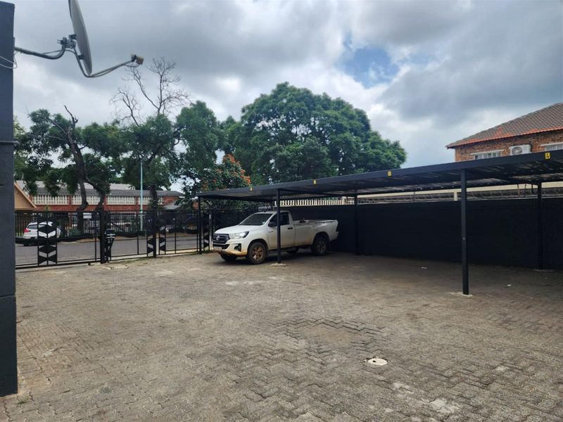 An outdoor paved parking area featuring a covered carport structure, secure perimeter fencing, and gated access.