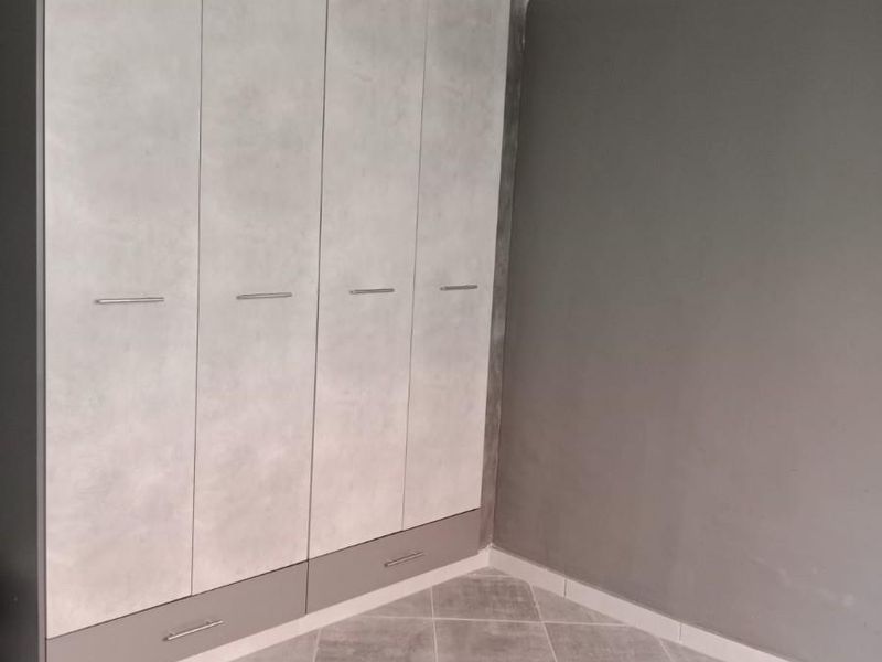 A clean, modern bedroom featuring a large built-in wardrobe with grey and stone-effect finishes, complemented by neutral tiled flooring and a glossy ceiling.