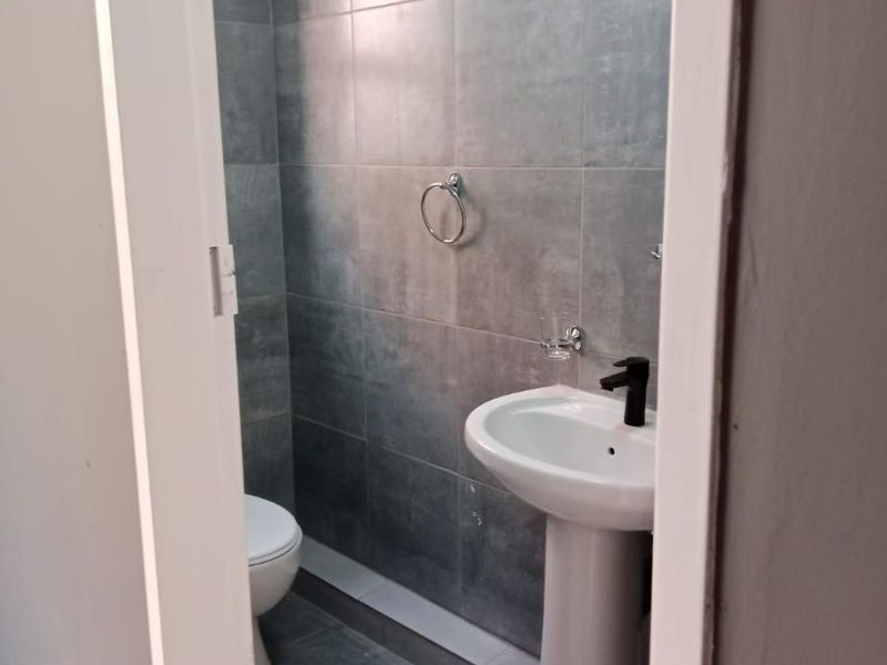 A compact, modern bathroom featuring grey tiled walls and flooring, a white pedestal basin with a black mixer tap, and a toilet.