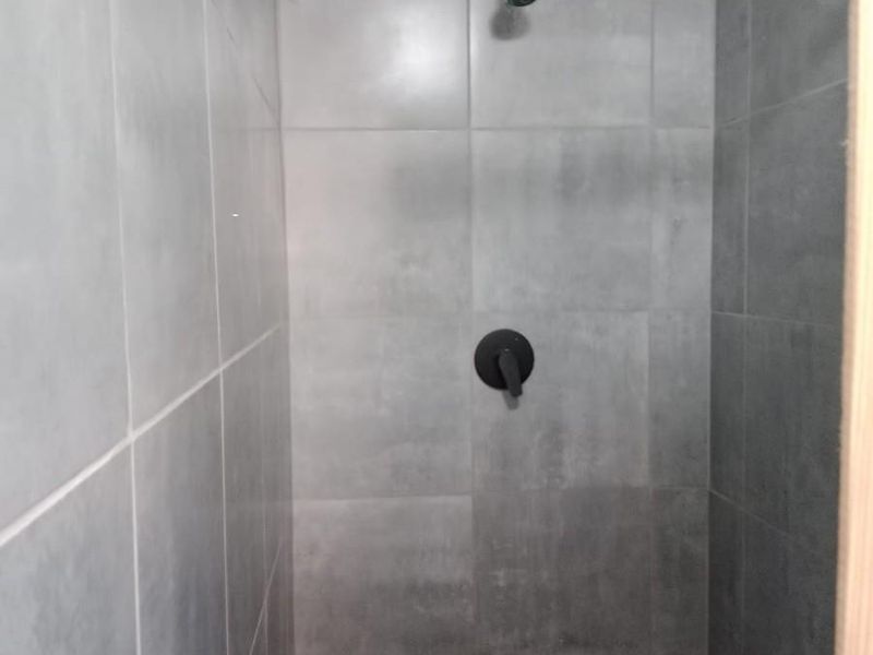 A modern shower enclosure featuring grey wall and floor tiles, a black shower mixer, and a standard shower head.