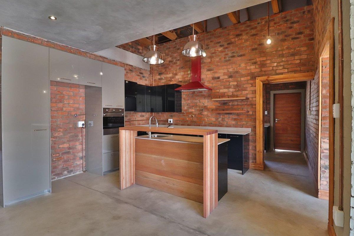 Modern industrial-style kitchen featuring exposed brick walls, concrete floors, timber ceiling beams, and a central island with integrated cabinetry and appliances.