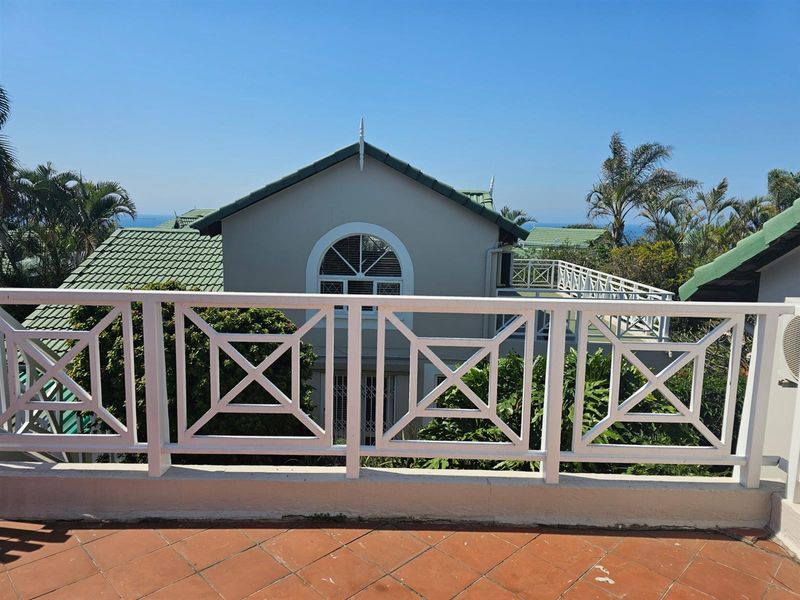 A spacious outdoor balcony featuring white balustrade railings, terracotta tiled flooring, and views of the surrounding property and coastal landscape.