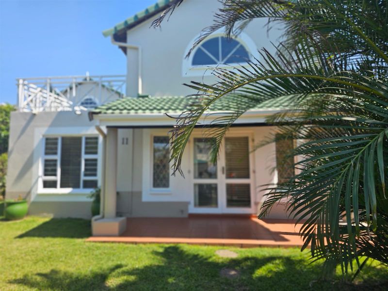 Rear view of the townhouse featuring a covered tiled patio, upper-level balcony, and well-maintained garden area.