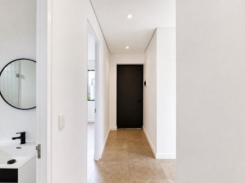 A clean, modern hallway featuring neutral tiled flooring, recessed downlighting, and a dark feature door, leading to adjacent living spaces.