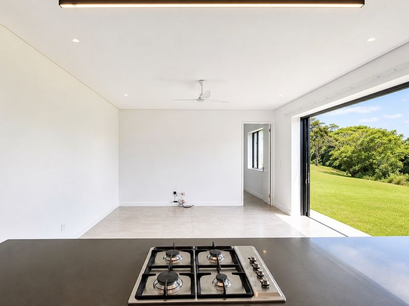 Modern open-plan living area featuring a sleek kitchen island with gas hob, tiled floors, and expansive sliding doors opening to a lush green landscape.