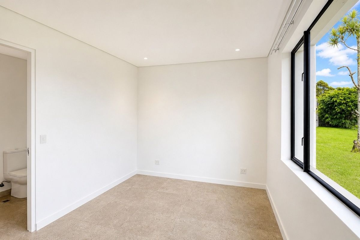 A bright, empty bedroom featuring neutral tiled flooring, recessed lighting, and a large window with views of the garden, including an attached en-suite bathroom.