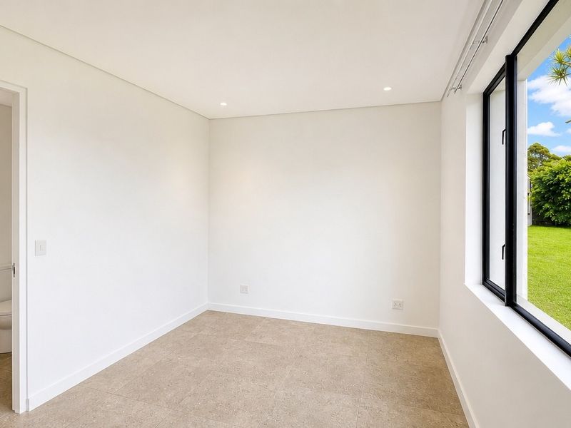 A bright, empty bedroom featuring neutral tiled flooring, recessed lighting, and a large window with views of the garden, including an attached en-suite bathroom.