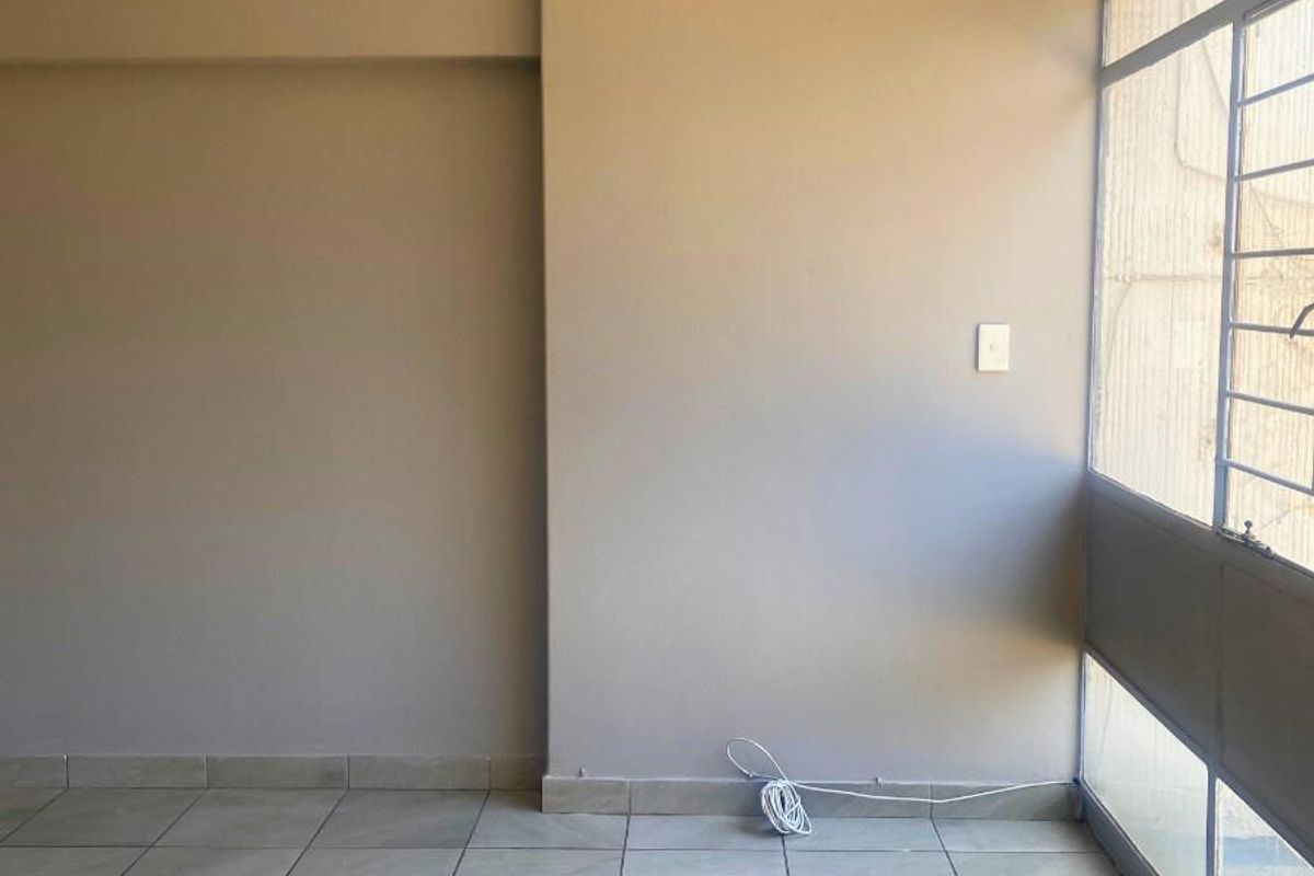An empty room featuring neutral wall tones, large windows with security bars, and light-colored floor tiles.