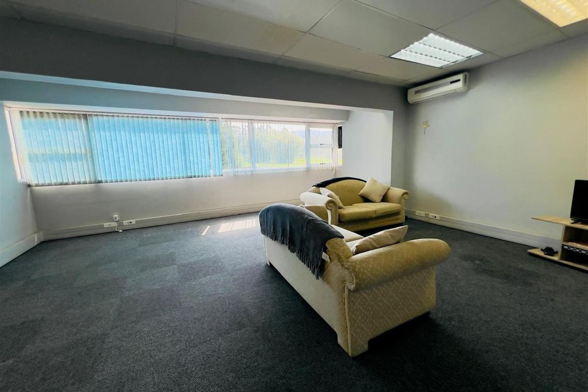 A spacious living area featuring dark carpeting, large windows with vertical blinds, and a wall-mounted air conditioning unit.