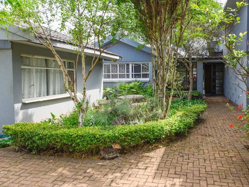 Front entrance area featuring a brick paved walkway, established garden beds, and mature trees leading to the main building entrance.