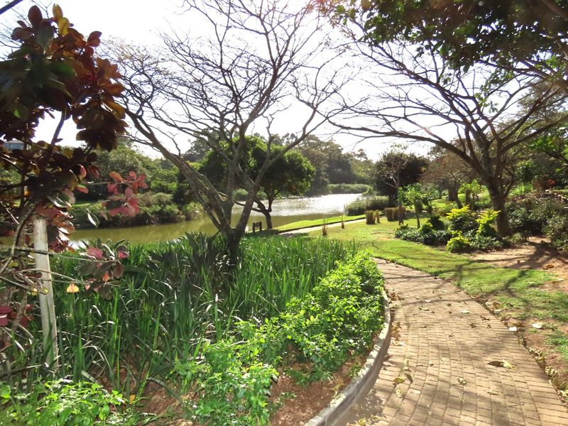 A scenic view of a landscaped garden featuring a paved walkway, mature trees, and a tranquil pond, highlighting the natural beauty of the property.
