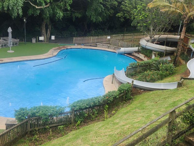 A well-maintained outdoor pool area featuring a large swimming pool, integrated water slide, and surrounding landscaped garden with artificial turf.