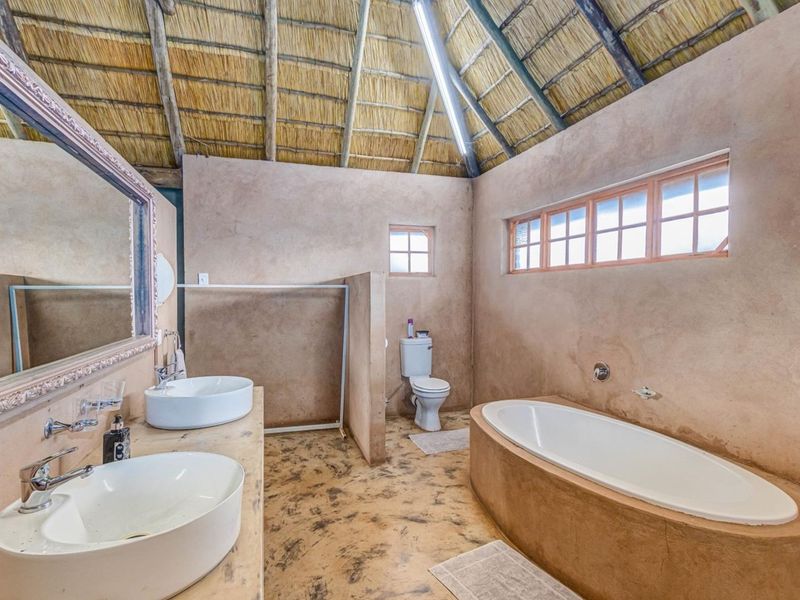 Spacious bathroom featuring a double vanity, built-in bathtub, toilet, and unique exposed thatch roof with rustic concrete wall and floor finishes.