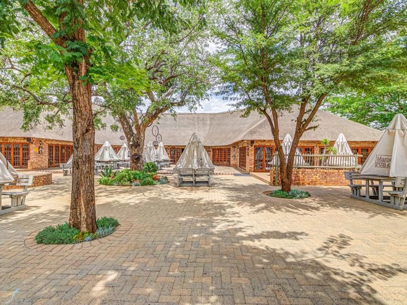 Spacious outdoor courtyard featuring brick paving, mature trees, and a large thatched-roof commercial building with multiple access points.