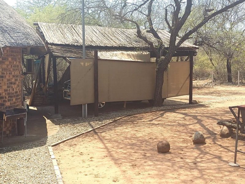Rear exterior view of a rustic property featuring a thatched roof, brick construction, and a covered carport area set within a natural bushveld environment.