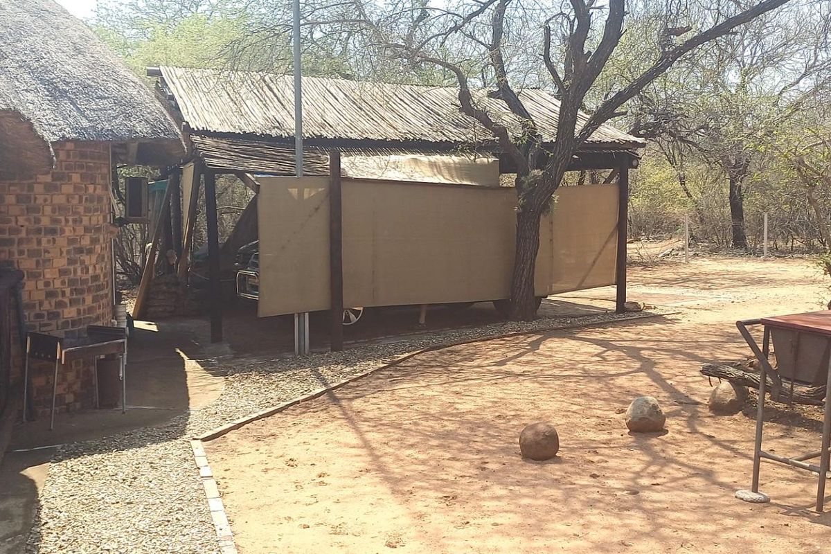 Rear exterior view of a rustic property featuring a thatched roof, brick construction, and a covered carport area set within a natural bushveld environment.