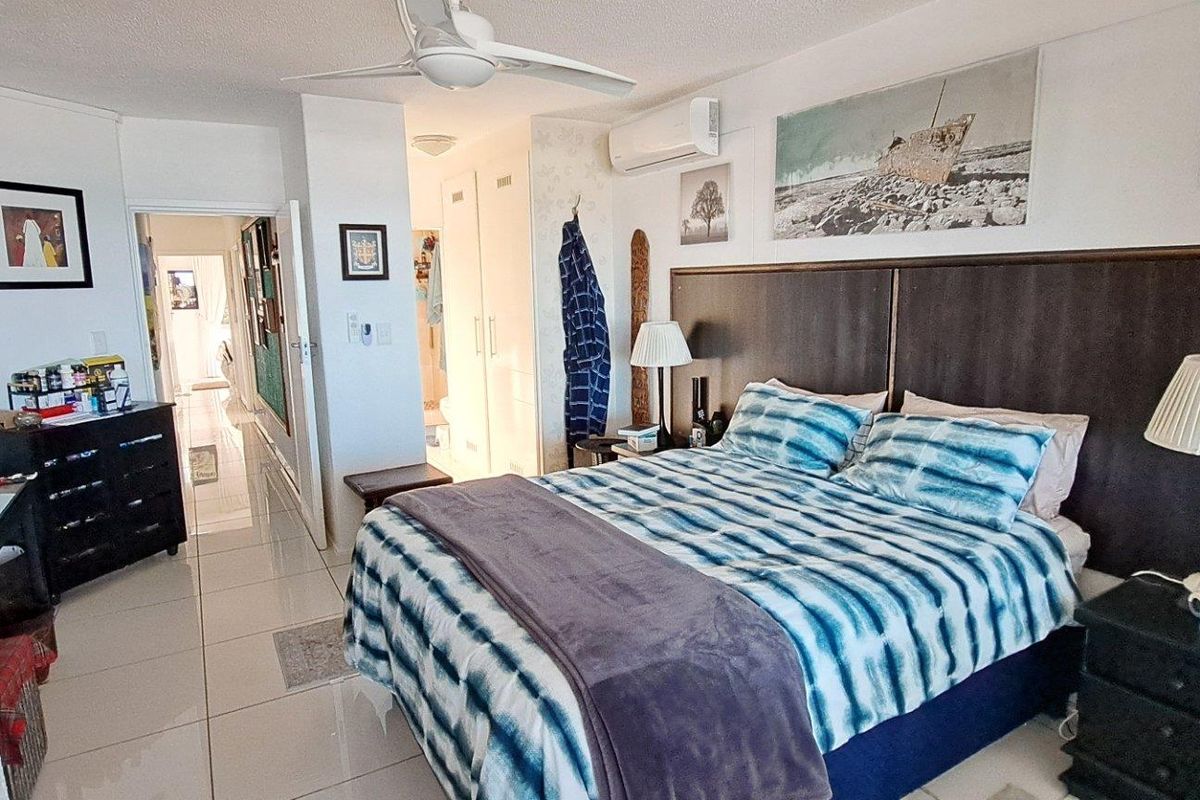 A well-lit bedroom featuring tiled flooring, a ceiling fan, and a wall-mounted air conditioning unit, with an adjoining hallway and built-in storage.
