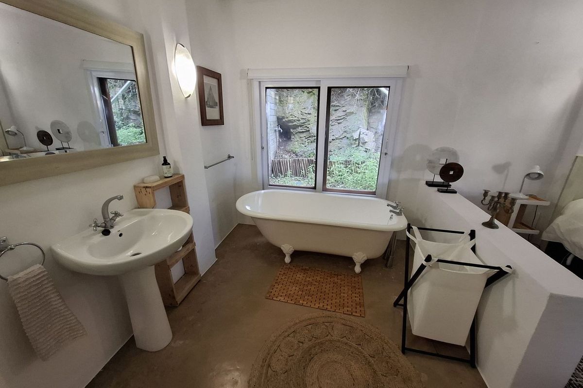 A bathroom featuring a classic clawfoot bathtub, pedestal sink, and polished concrete flooring with a view of the natural rock face outside.