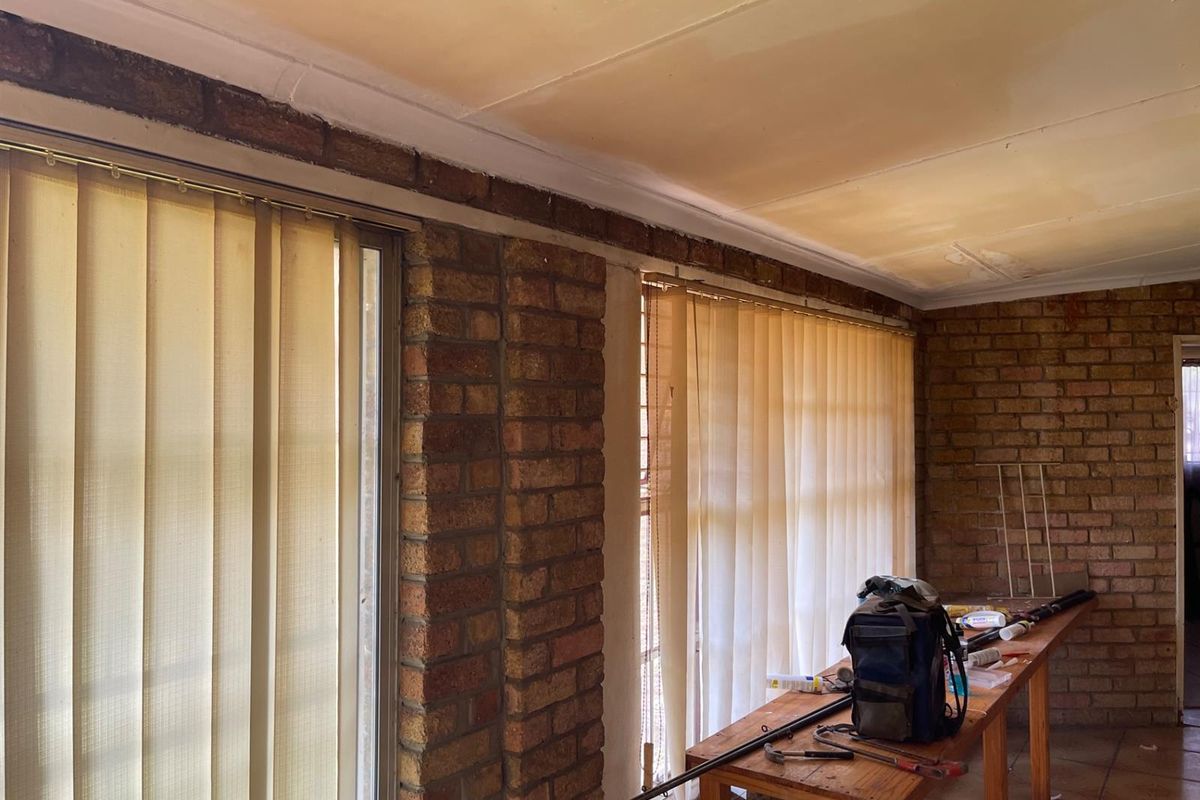 A living area featuring exposed brick walls, tiled flooring, and large windows with vertical blinds, currently undergoing maintenance or renovation.