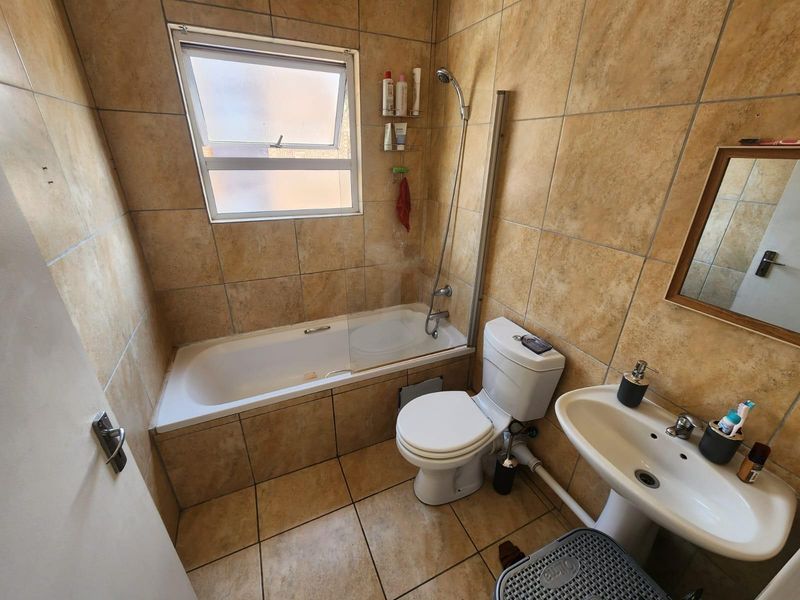 A functional bathroom featuring a bathtub with a shower mixer, glass screen, pedestal basin, and toilet, finished with neutral floor-to-ceiling wall tiles.