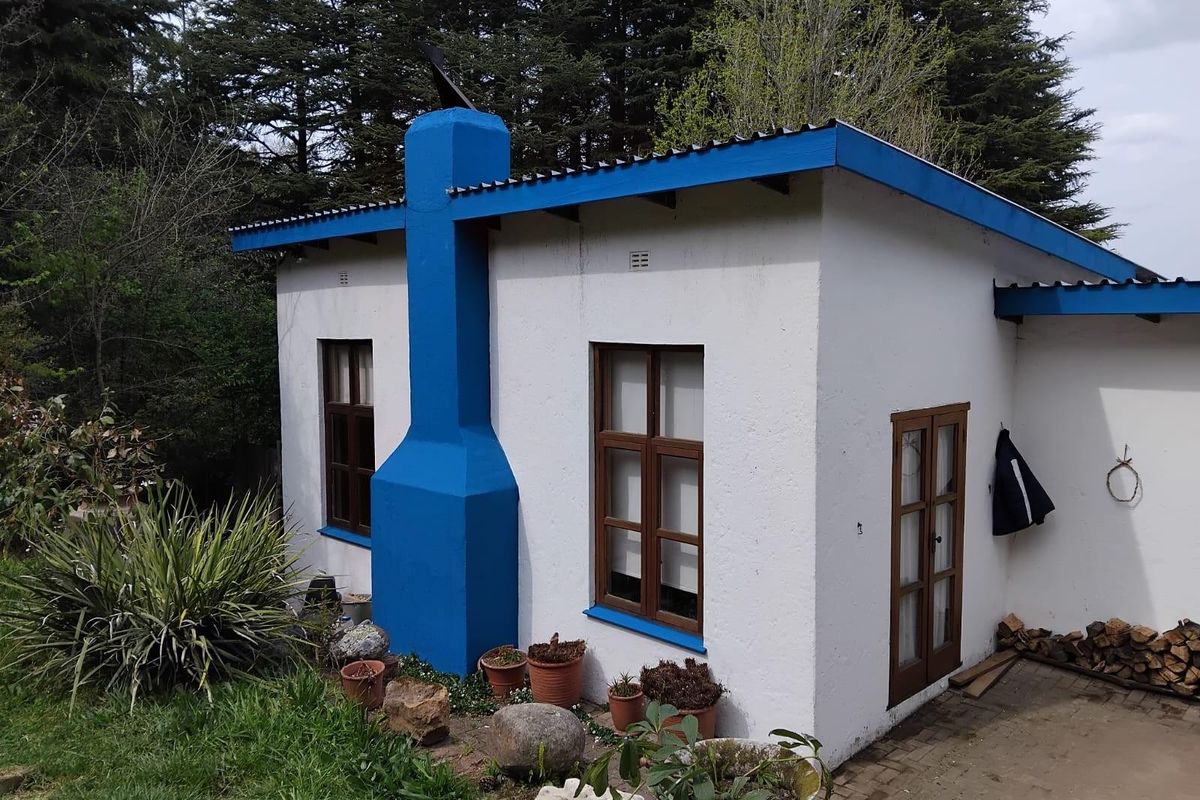 Exterior view of a small detached structure featuring a prominent blue chimney, white rendered walls, and timber-framed windows set in a garden.