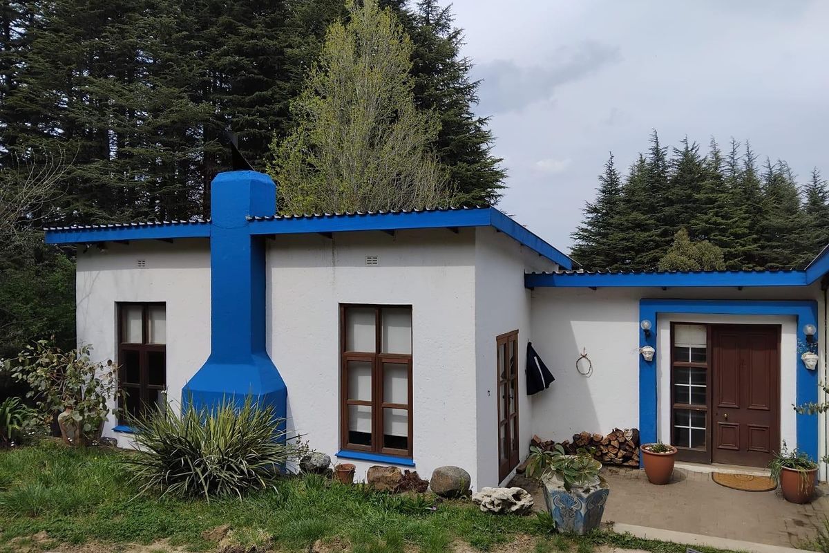 A charming cottage-style exterior featuring a prominent blue chimney, white rendered walls, and a covered entrance area surrounded by established trees.