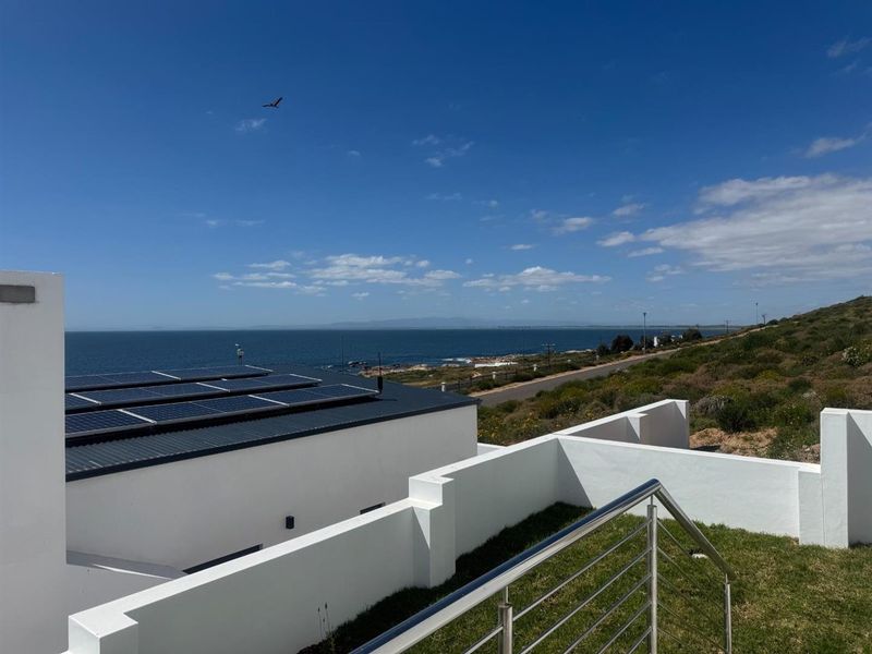 Elevated view of the property exterior featuring a modern roof with solar panels, white rendered walls, and a stainless steel balustrade overlooking the coastline.