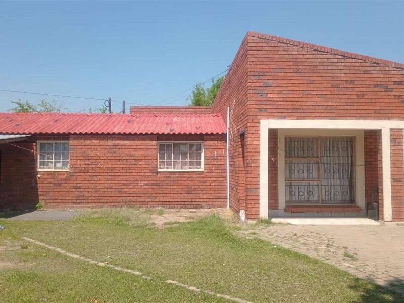 A single-story face brick house featuring a corrugated metal roof, security gates, and a paved driveway area with an adjacent lawn.