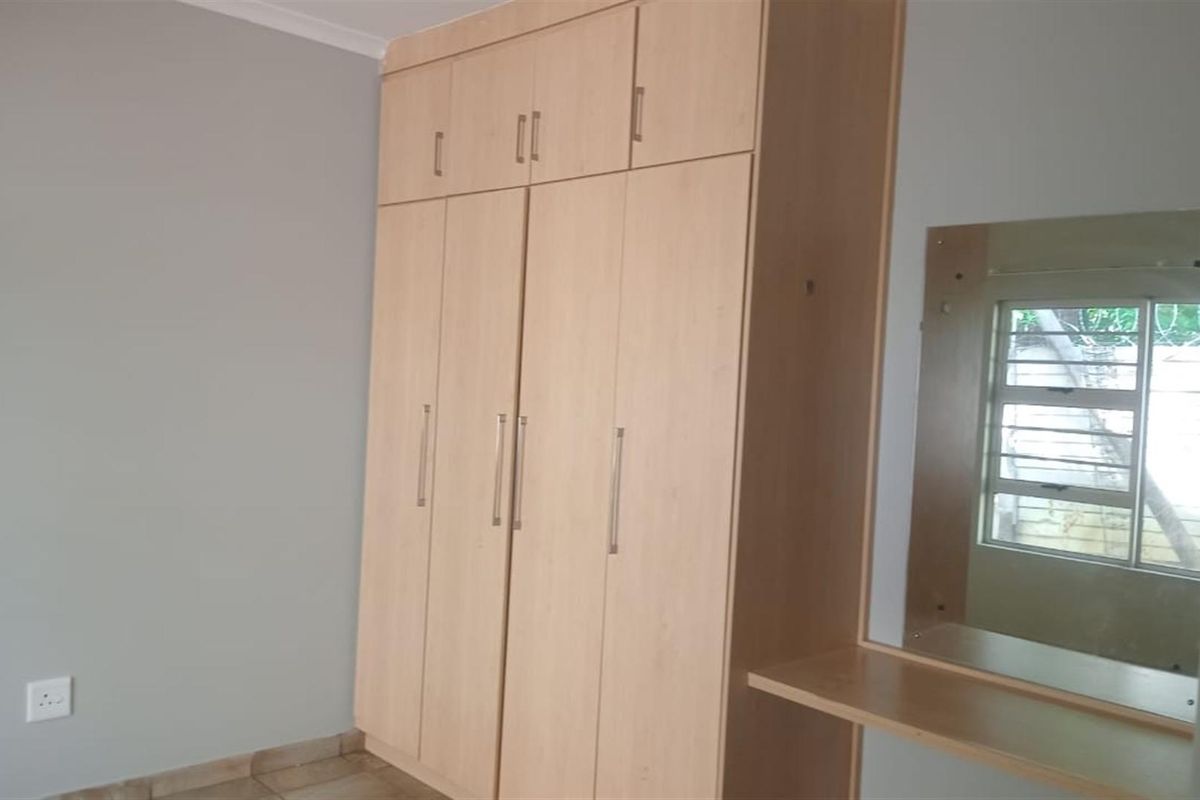A clean bedroom featuring light wood-finish built-in cupboards with an integrated vanity mirror and shelf, complemented by neutral wall tones and tiled flooring.