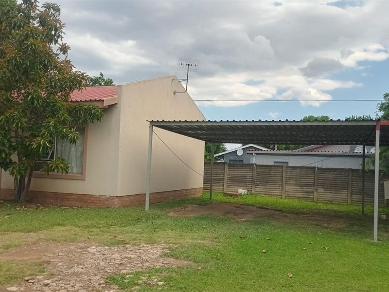 Rear exterior view featuring a large lawn, a metal carport structure, and a precast concrete boundary wall.