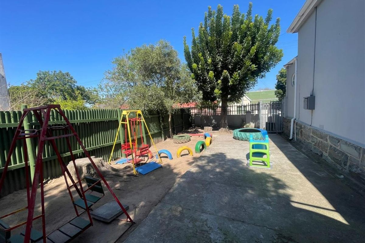 A rear garden area featuring a concrete patio, sandy play zone, and established trees, enclosed by perimeter fencing.