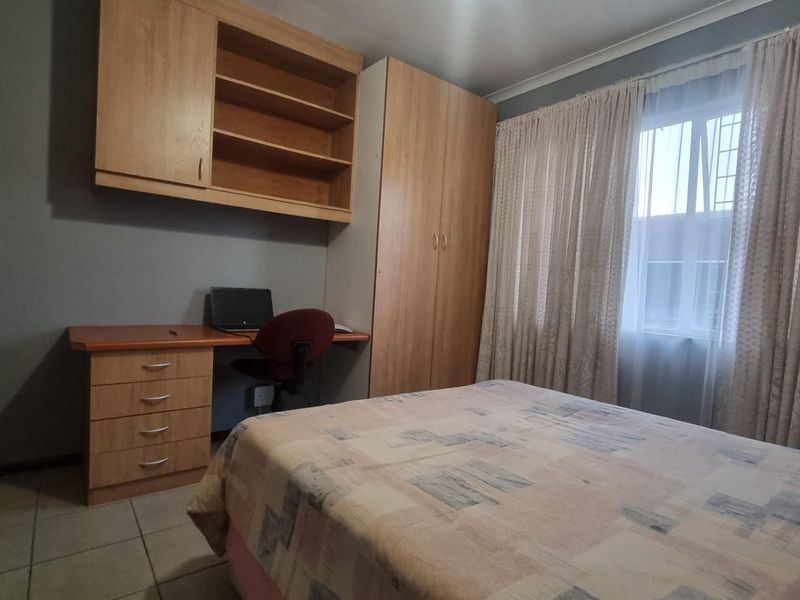 A functional bedroom featuring built-in wooden cabinetry, a desk area, tiled flooring, and large windows with curtains.