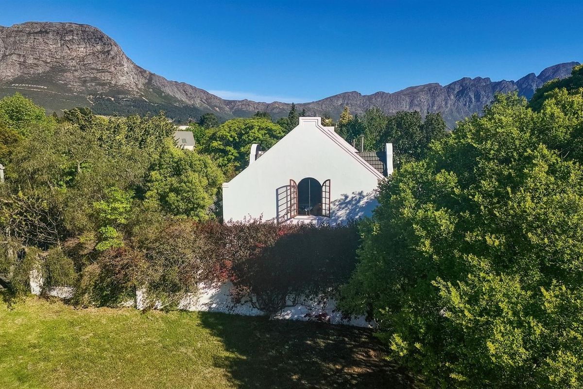 A scenic view of the property's rear elevation, featuring traditional Cape Dutch architecture nestled among lush trees with a dramatic mountain backdrop.