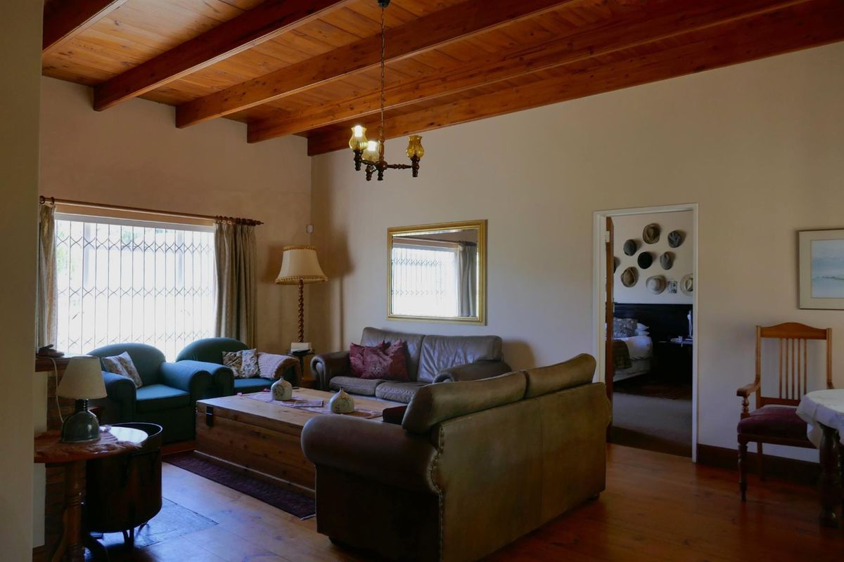 A cozy living room featuring exposed timber beam ceilings, warm wooden flooring, and a large window with security bars, leading into an adjacent bedroom.