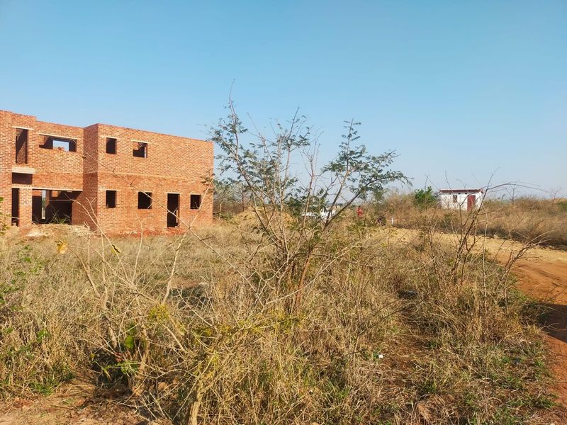 A view of a residential plot featuring an unfinished brick building structure surrounded by dry, overgrown vegetation and a dirt access road.