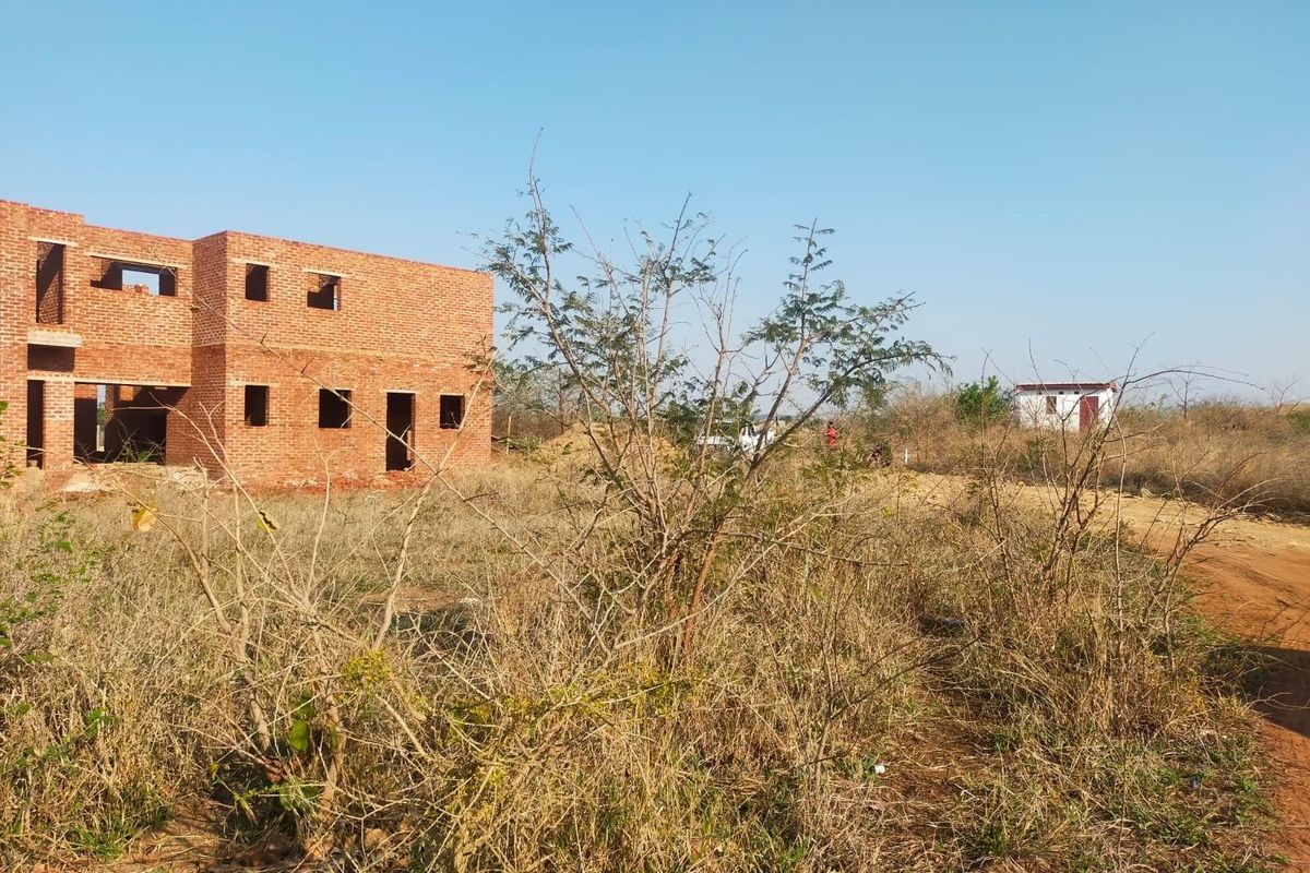 A view of a residential plot featuring an unfinished brick building structure surrounded by dry, overgrown vegetation and a dirt access road.