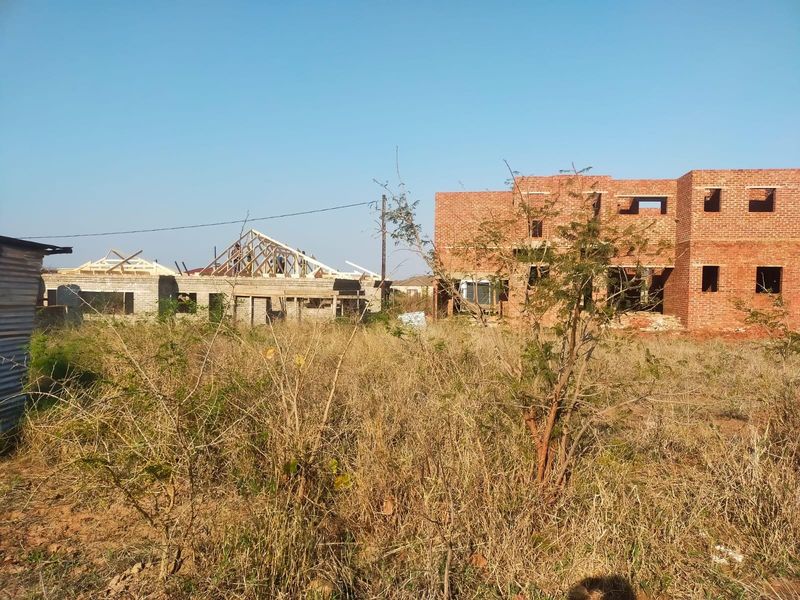 A view of a residential property under construction, featuring exposed brickwork and timber roof trusses on a plot with overgrown vegetation.