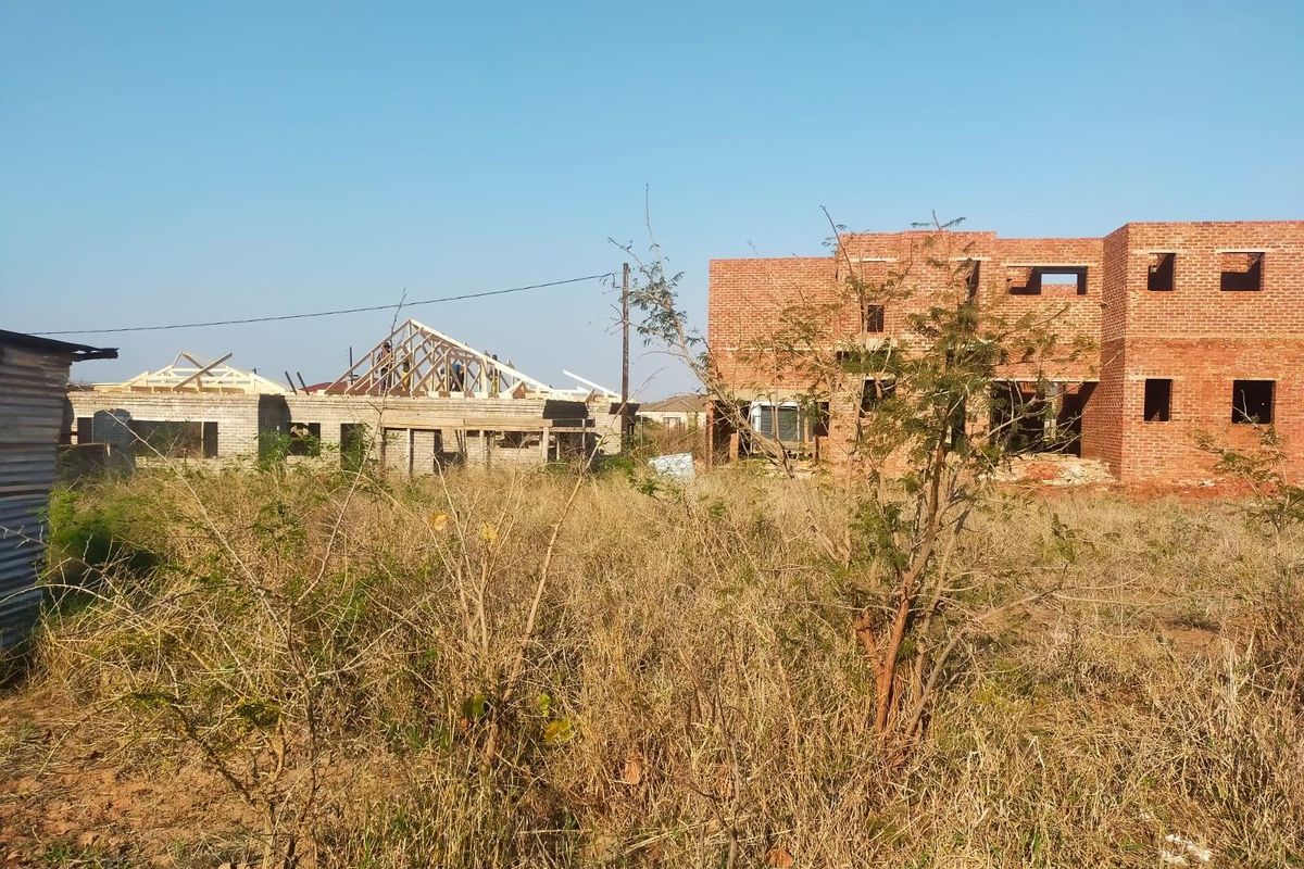 A view of a residential property under construction, featuring exposed brickwork and timber roof trusses on a plot with overgrown vegetation.