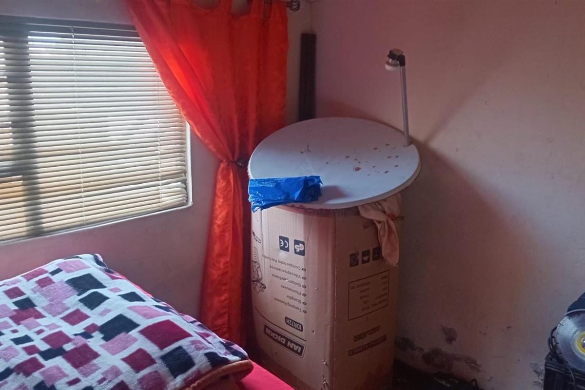 A bedroom interior featuring a window with fitted blinds and visible signs of wall surface deterioration near the floor.